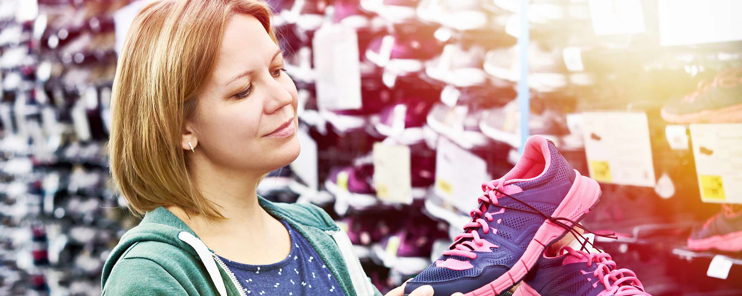 A woman at a store holds pink and purple running shoes as she thinks about whether buying them would cause her checking account to overdraft.