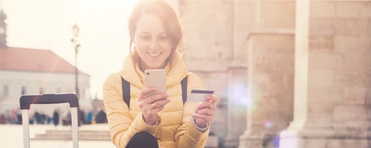 Woman smiles as she sits on stone steps and looks at her phone and debit card.