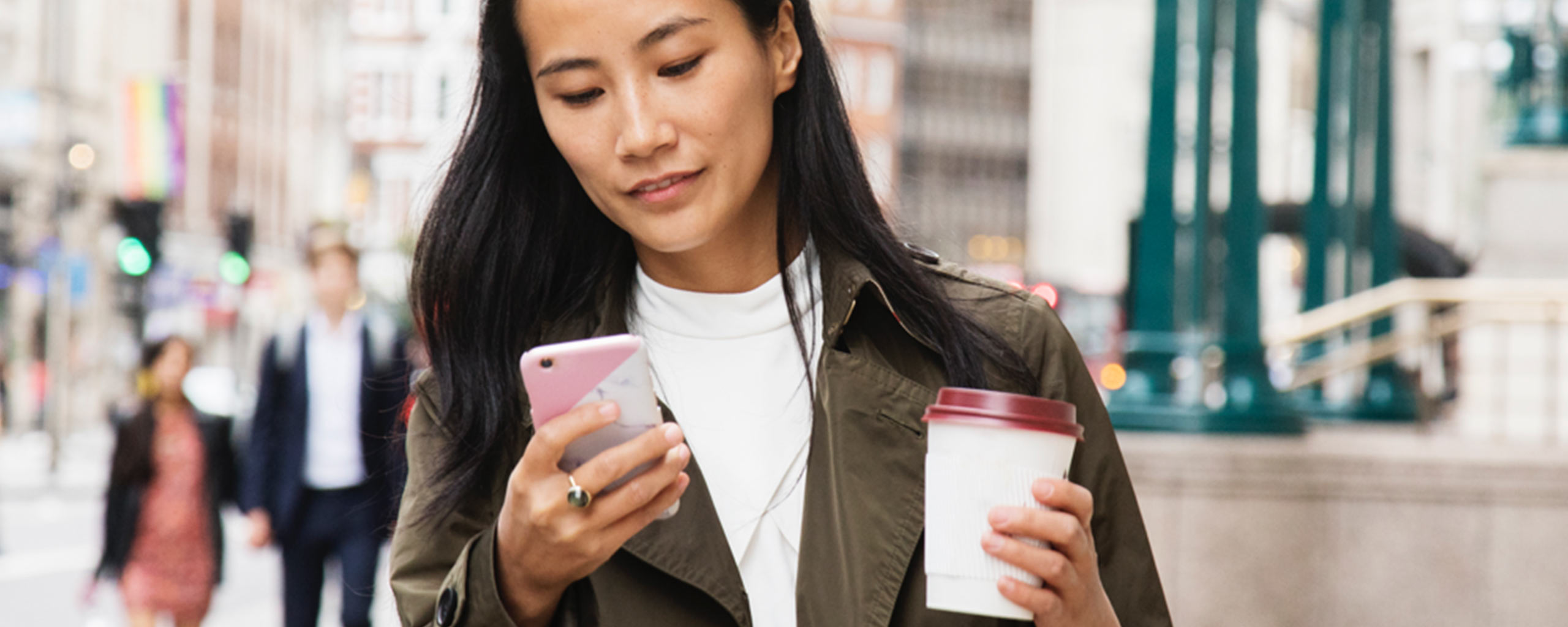 A woman with a coffee cup walks around a city and checks her phone to make sure she didn’t overdraft her checking account.