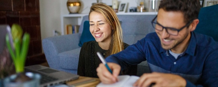 A woman with a laptop and a man with a notebook sit on the floor and smile after calculating the interest they’ve earned on their joint savings account.