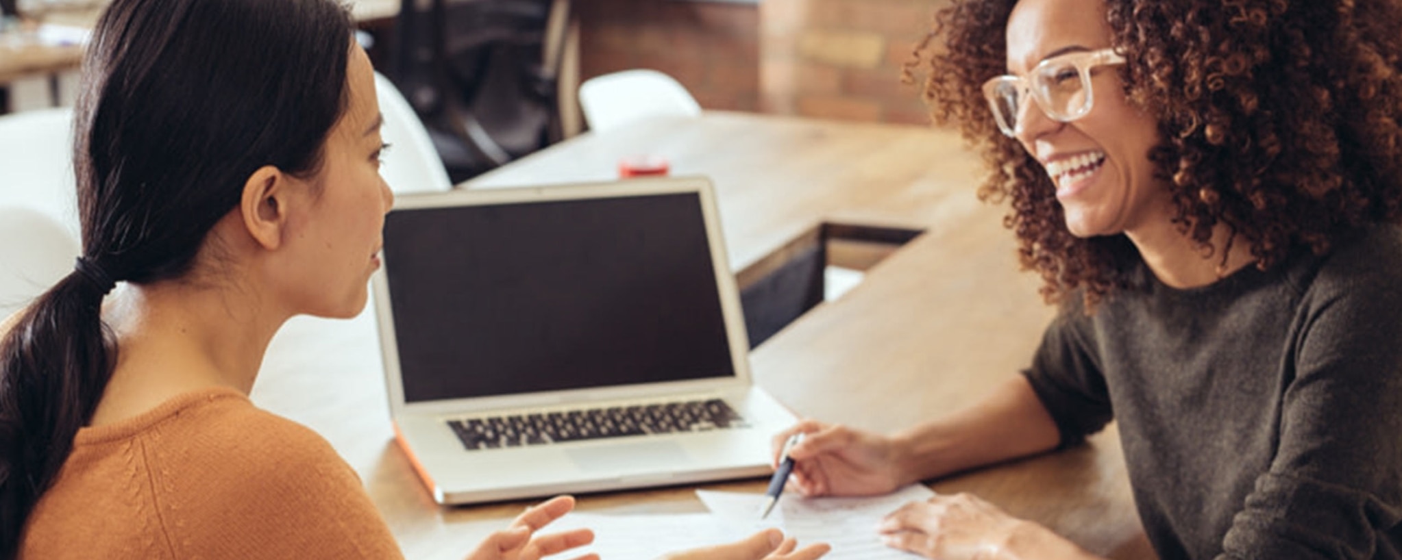 Two women sit together at a laptop as one explains to the other how secured and unsecured loans work.