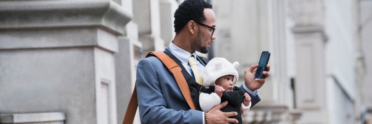A man in business attire carries a baby and checks his phone in front of a stone building.