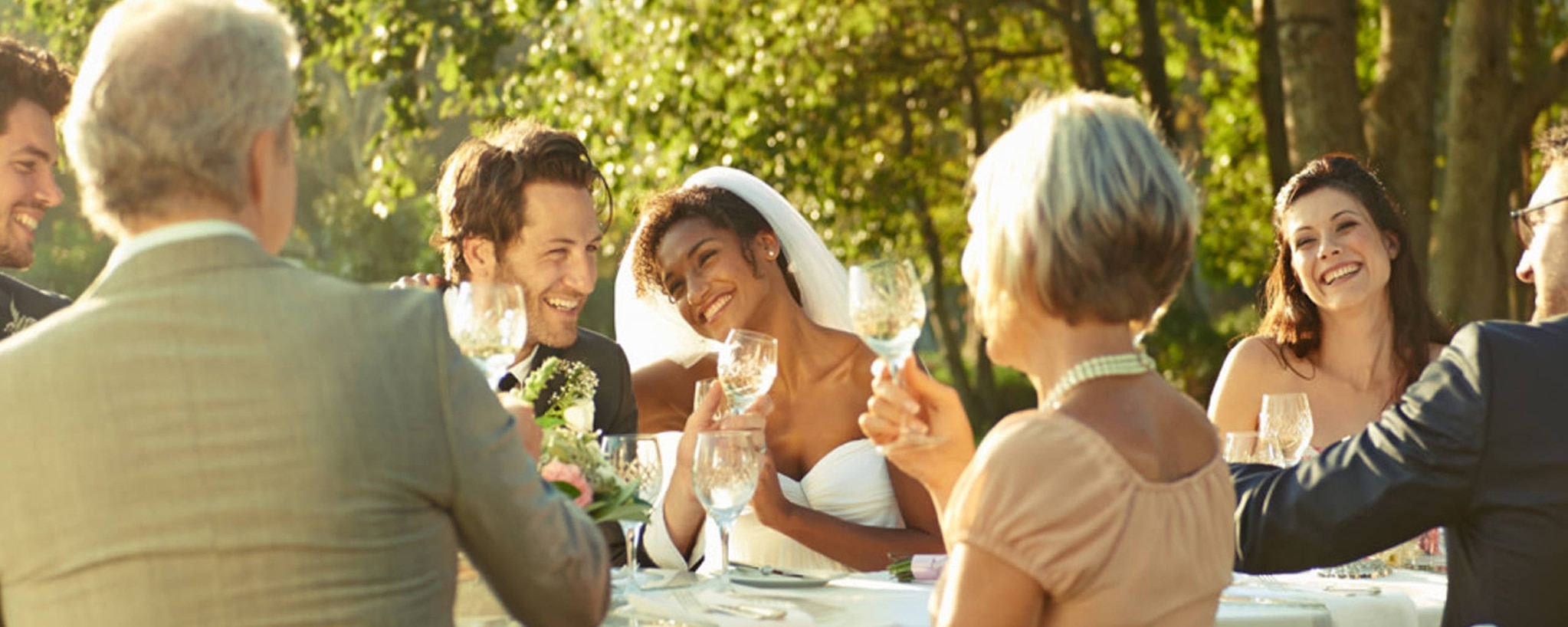 Bride and groom sit at an outdoor reception table and toast with their parents and guests.