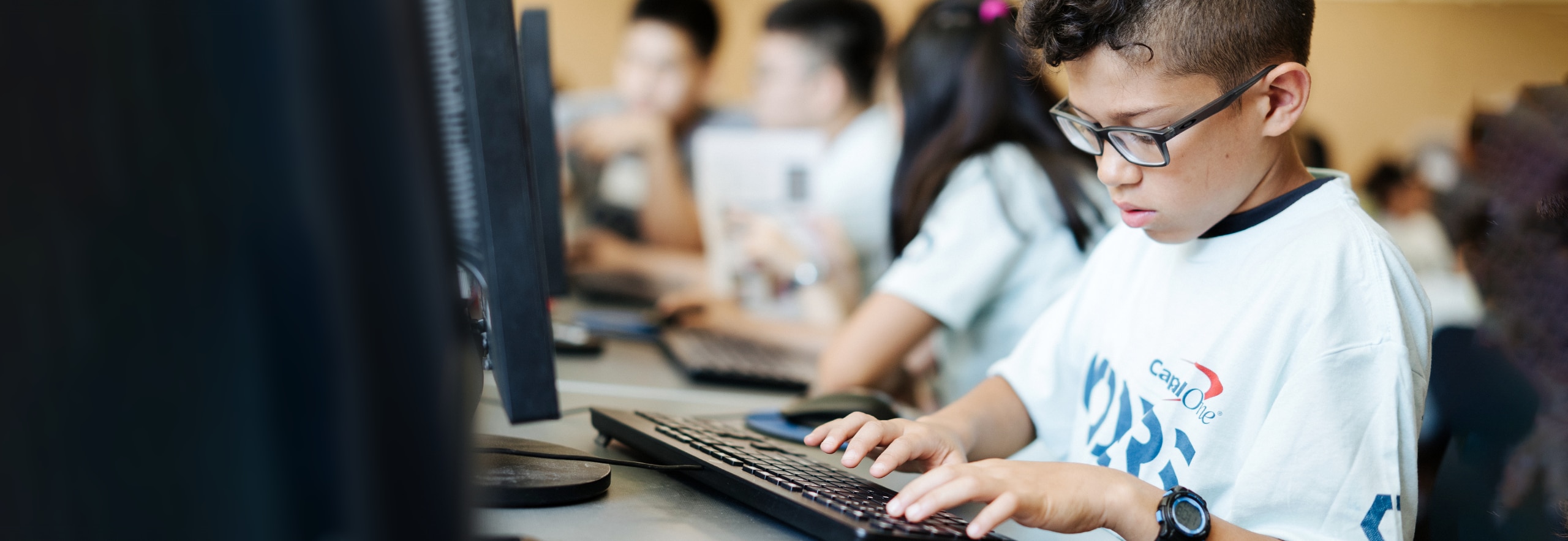 A student wears a Capital One Coders t-shirt and sits in a classroom with other Coders students, typing on a keyboard