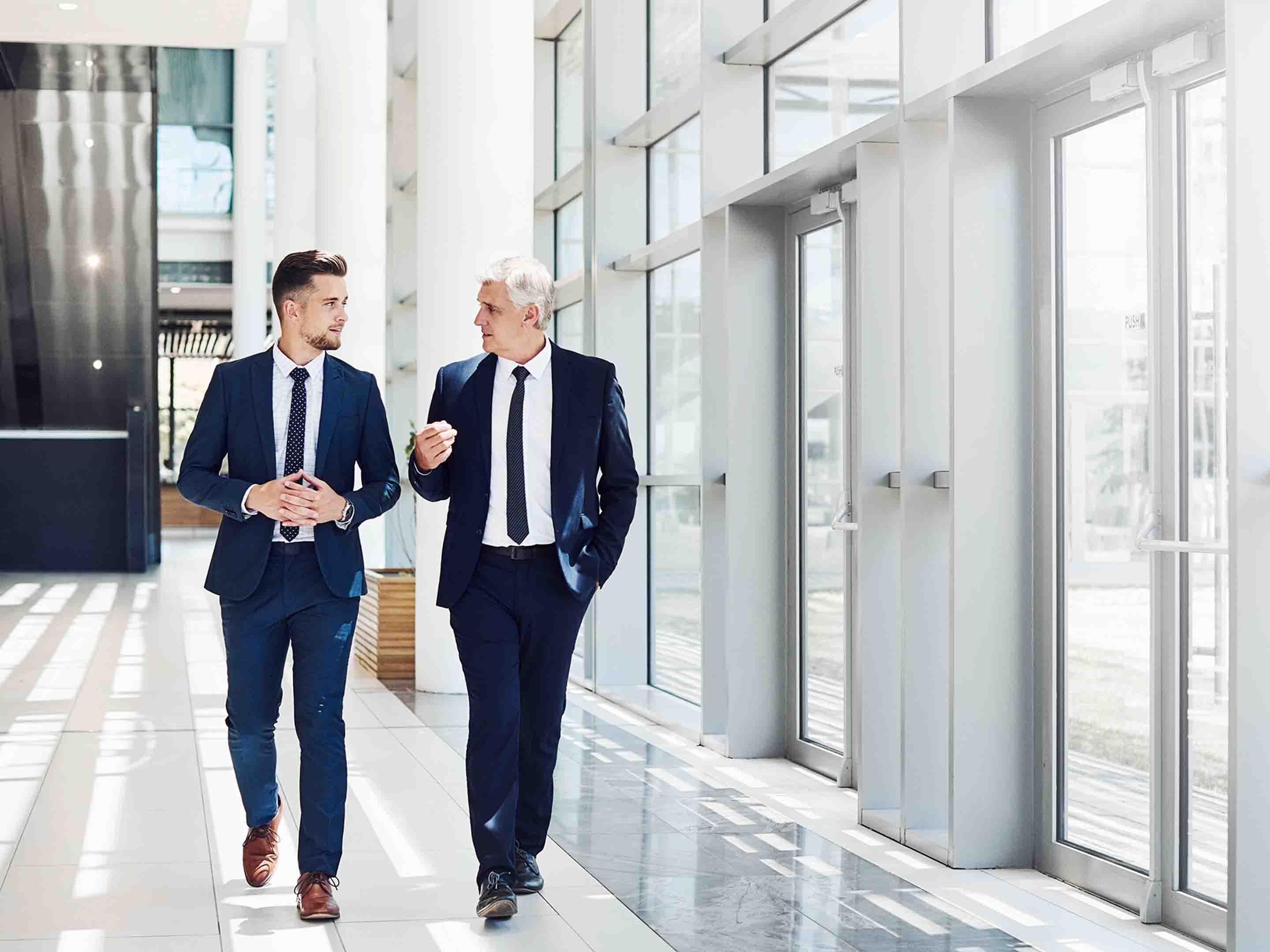 two men in suits walking and talking in a hallway in a corporate building