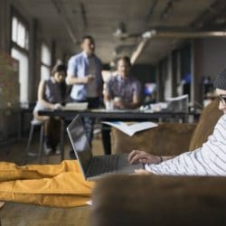 A man works on his laptop in an open office with coworkers grouped in the background.