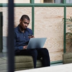Young man looks thoughtfully at his credit card as he sits with a laptop on his legs