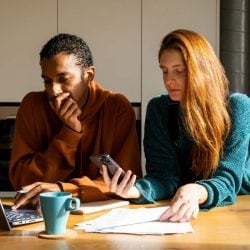 A couple sit together at a table looking thoughtfully at a laptop, phone, and paperwork.