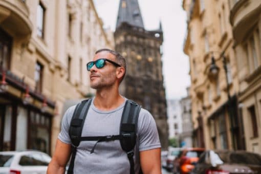 Man in sunglasses wearing backpack walks down European street.