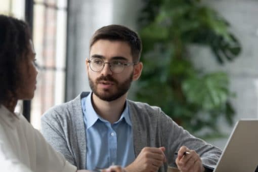 Man sits at desk in office with open laptop speaking to a woman next to him.
