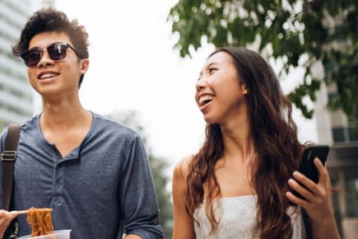 Two friends walking outside and smiling, one holding a phone and one eating.
