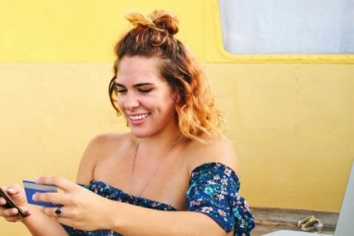 Smiling woman, standing next to a laptop, while looking at a credit card in one hand and mobile phone in another hand.