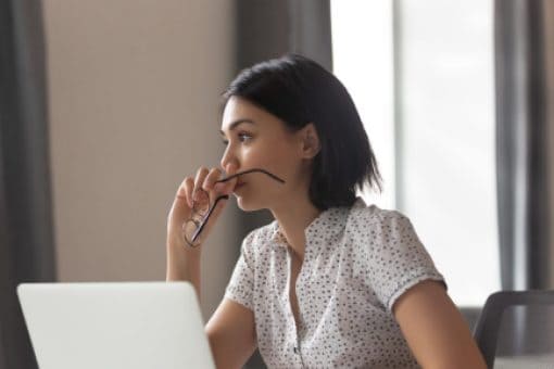 A woman with a contemplative expression sits in front of a laptop and stares into the distance.