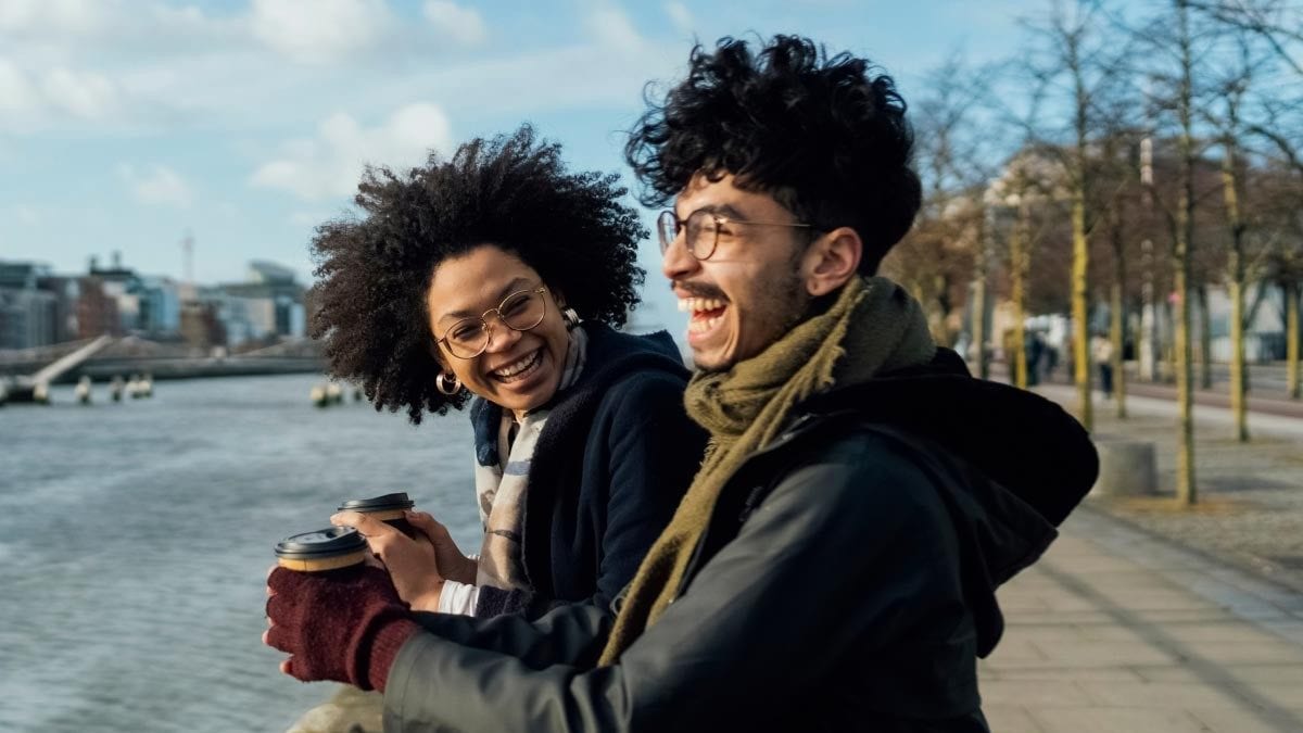A man and a woman holding coffee to-go cups laugh while looking out over a river.