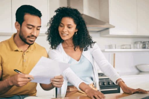 Two people discussing finances at a kitchen island while reviewing documents and using a laptop.