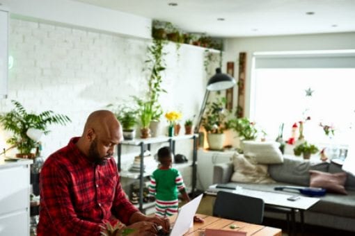 Father works on laptop as his son walks around the living room in the background.