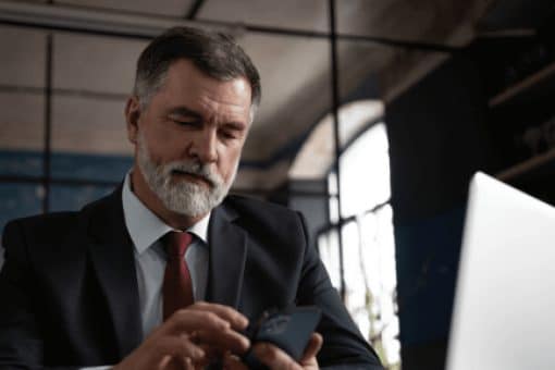 A man in a suit and tie uses his mobile phone and laptop in his office.