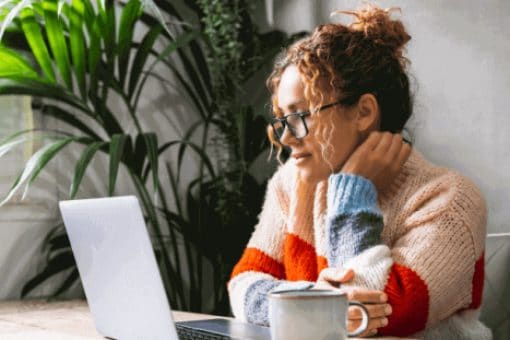 Person wearing glasses working on a laptop at home with a coffee mug nearby.