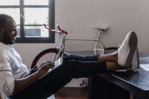 Happy man has his feet up on his desk as he leans back in his chair and looks at his laptop in his lap.