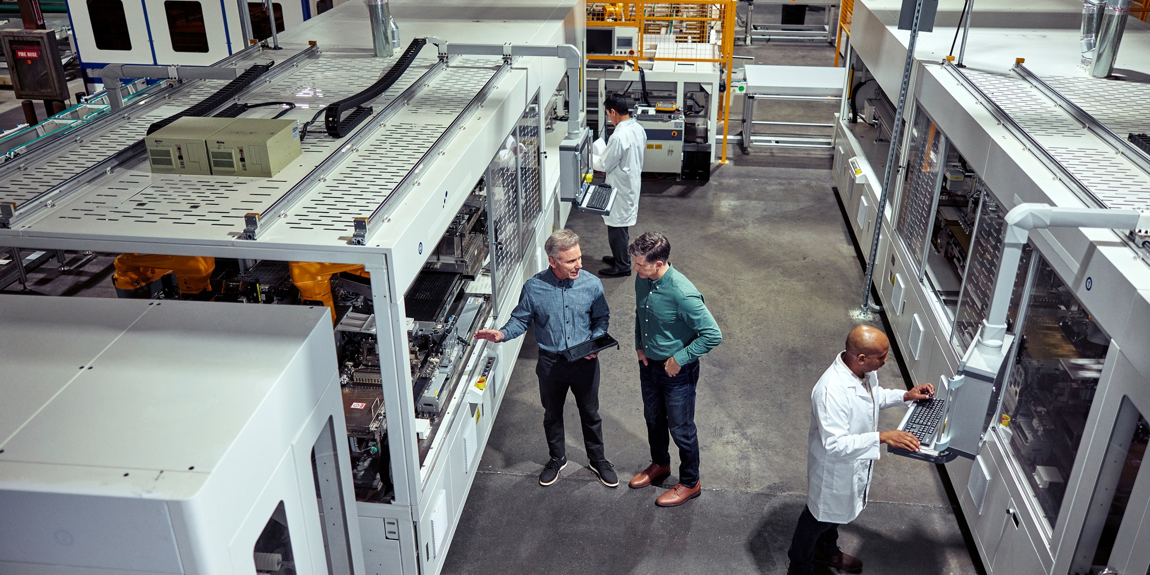 Two men talk beside large machinery. There are others in the room who are wearing white lab coats.