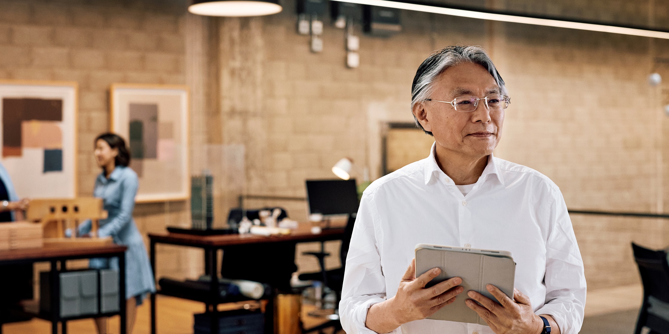 A man in a white button-down shirt stands in an office holding a digital tablet.