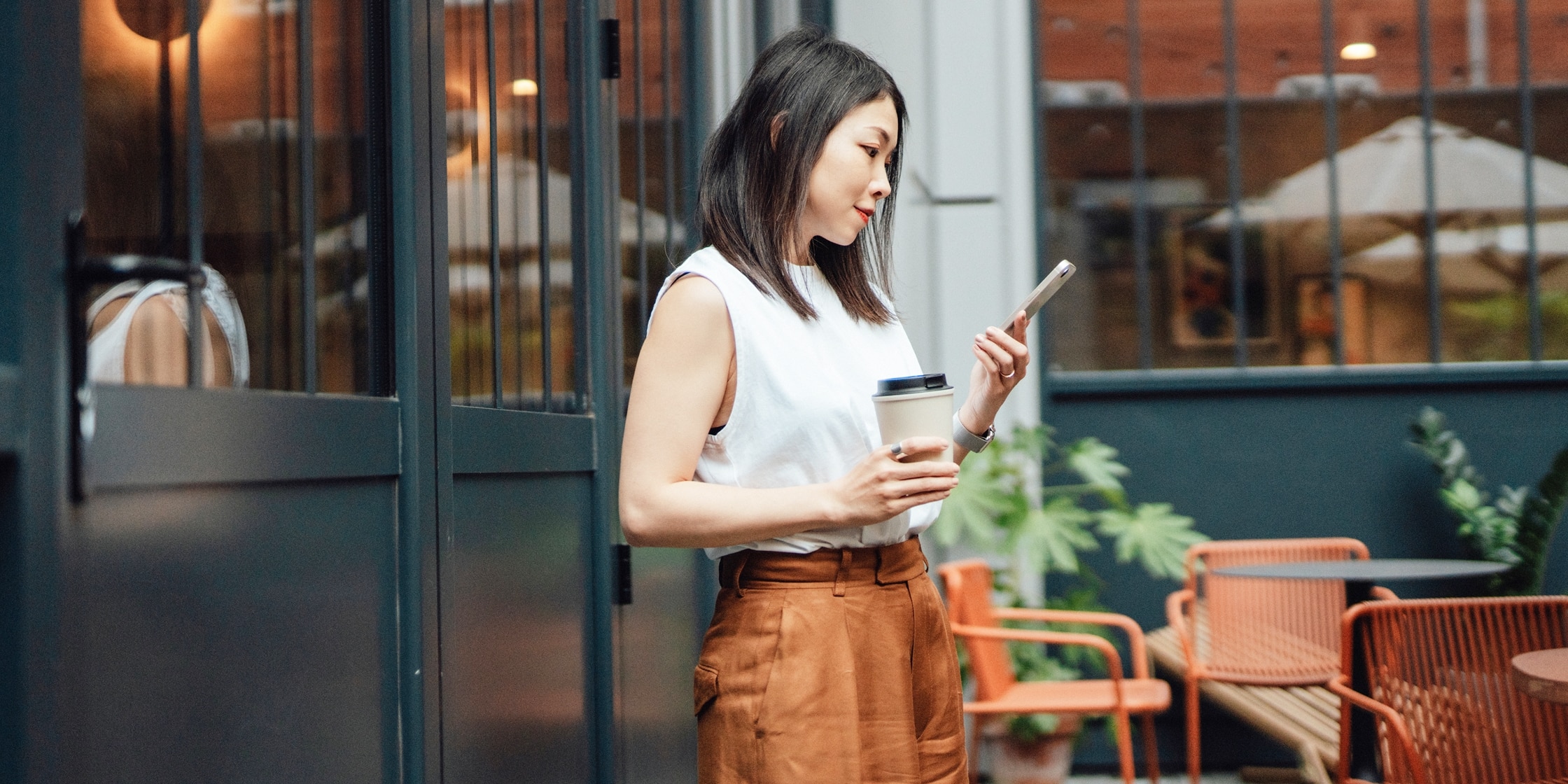 A woman stands on a patio holding a to-go coffee cup and looking at her phone.