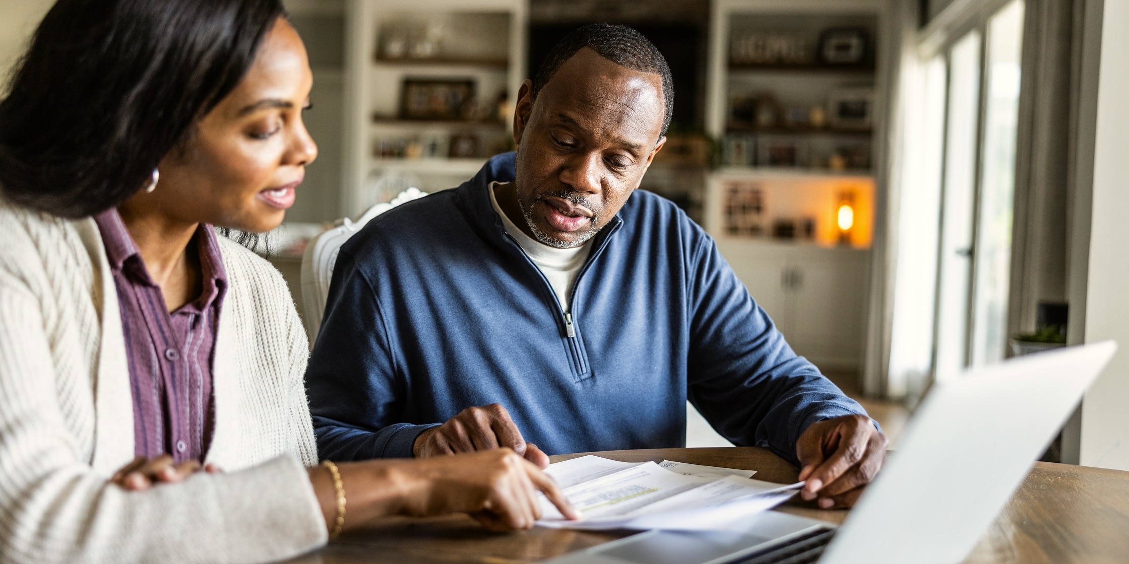A man and woman look over paperwork together while seated at their kitchen table.