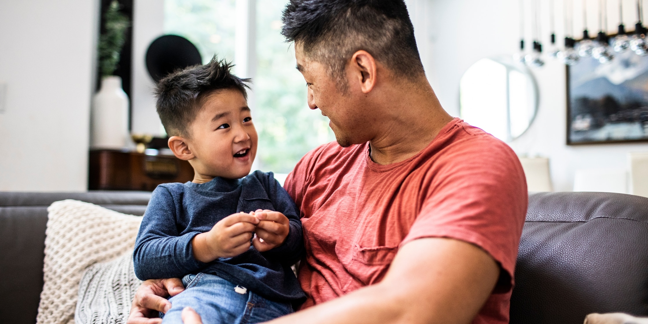A young boy sits in his father’s lap on their living room couch.