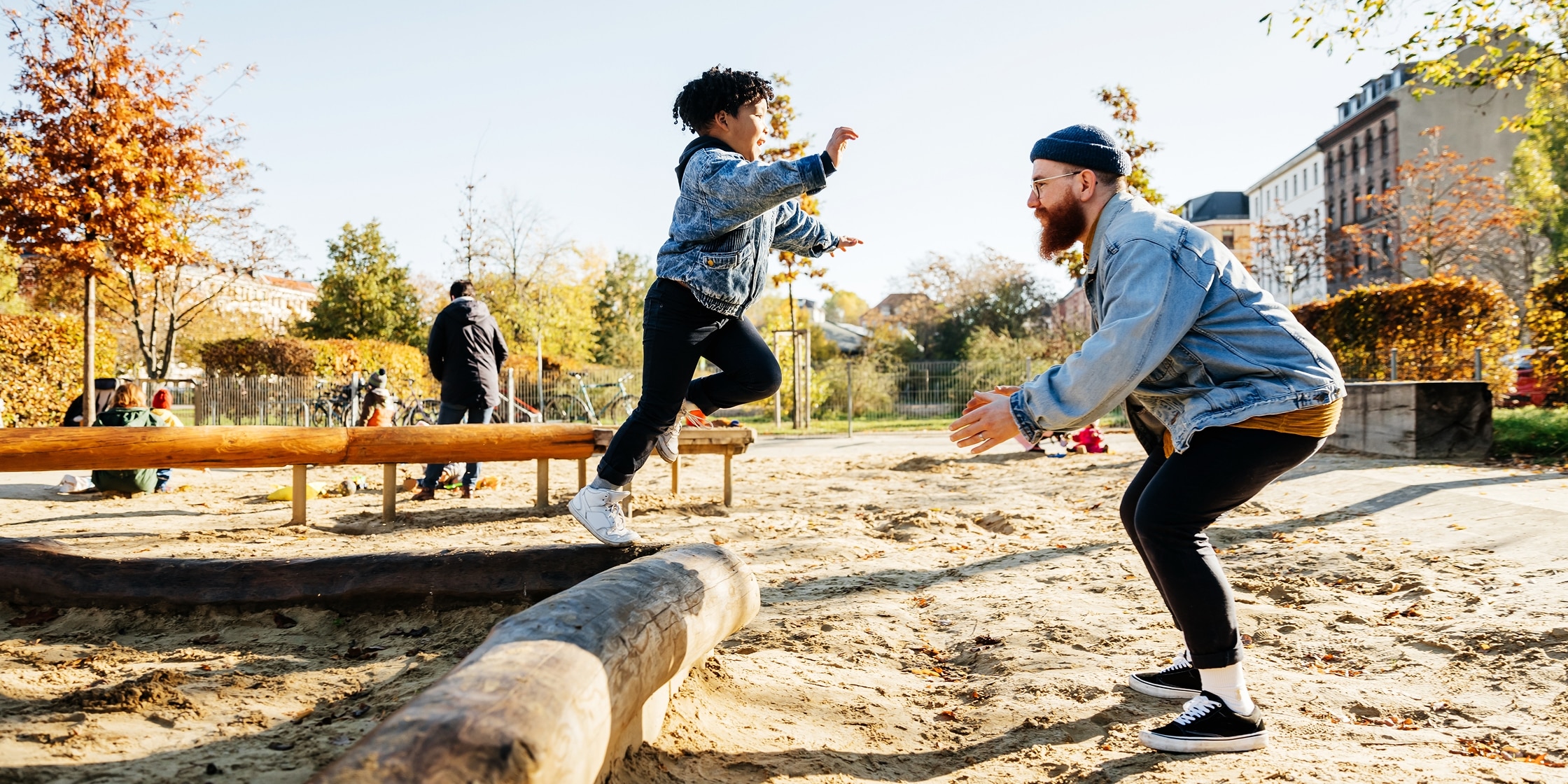 A young boy leaps off playground equipment while his father stands ready to catch him.