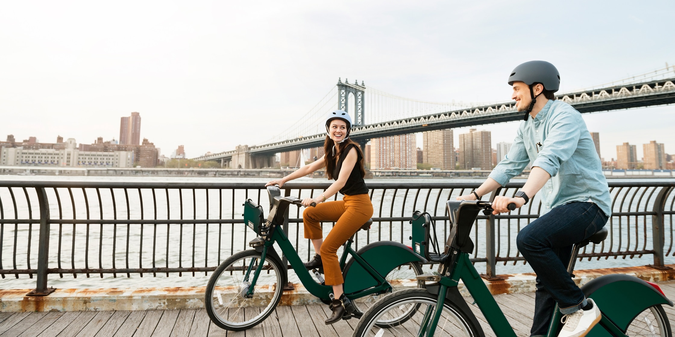 A man and woman ride side-by-side on electric bikes across a wooden bridge. In the background is a larger bridge and cityscape.