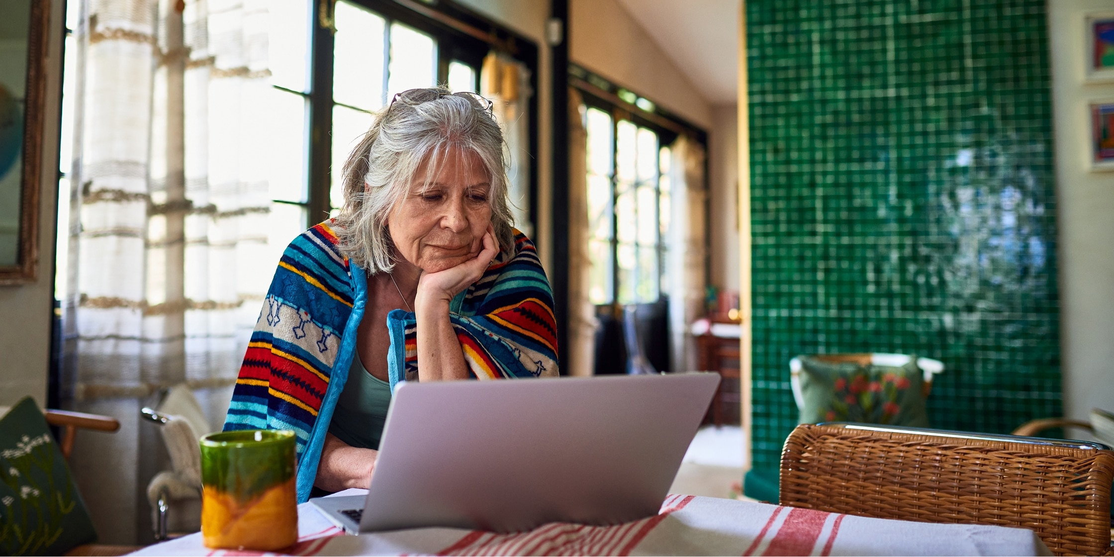 A senior woman scrolls on her laptop while seated at her kitchen table.