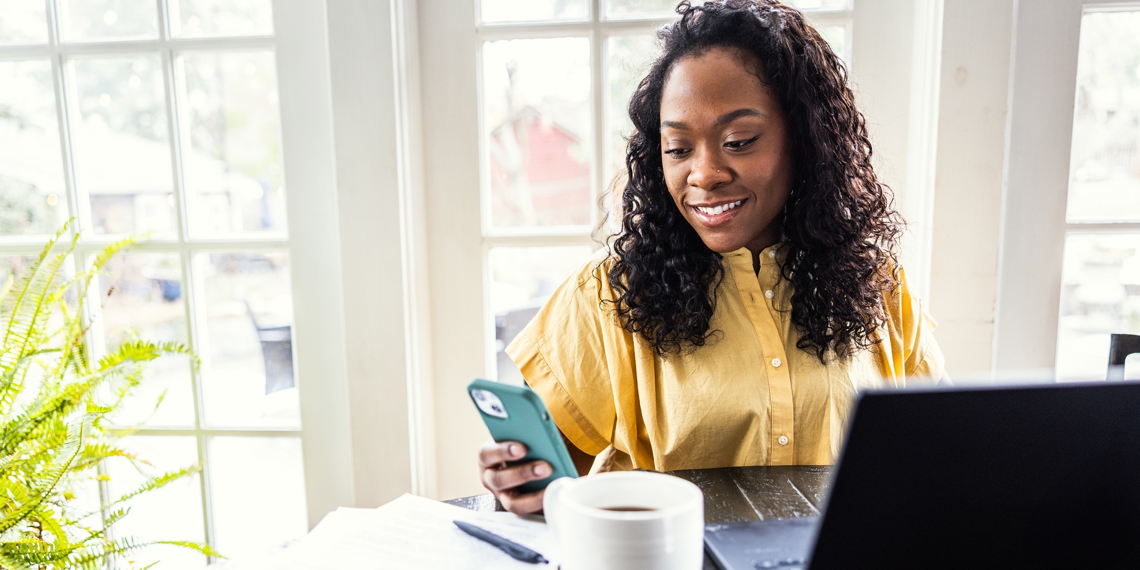 A woman sits at her kitchen table, looking at her phone while her laptop and coffee cup lay on the table.