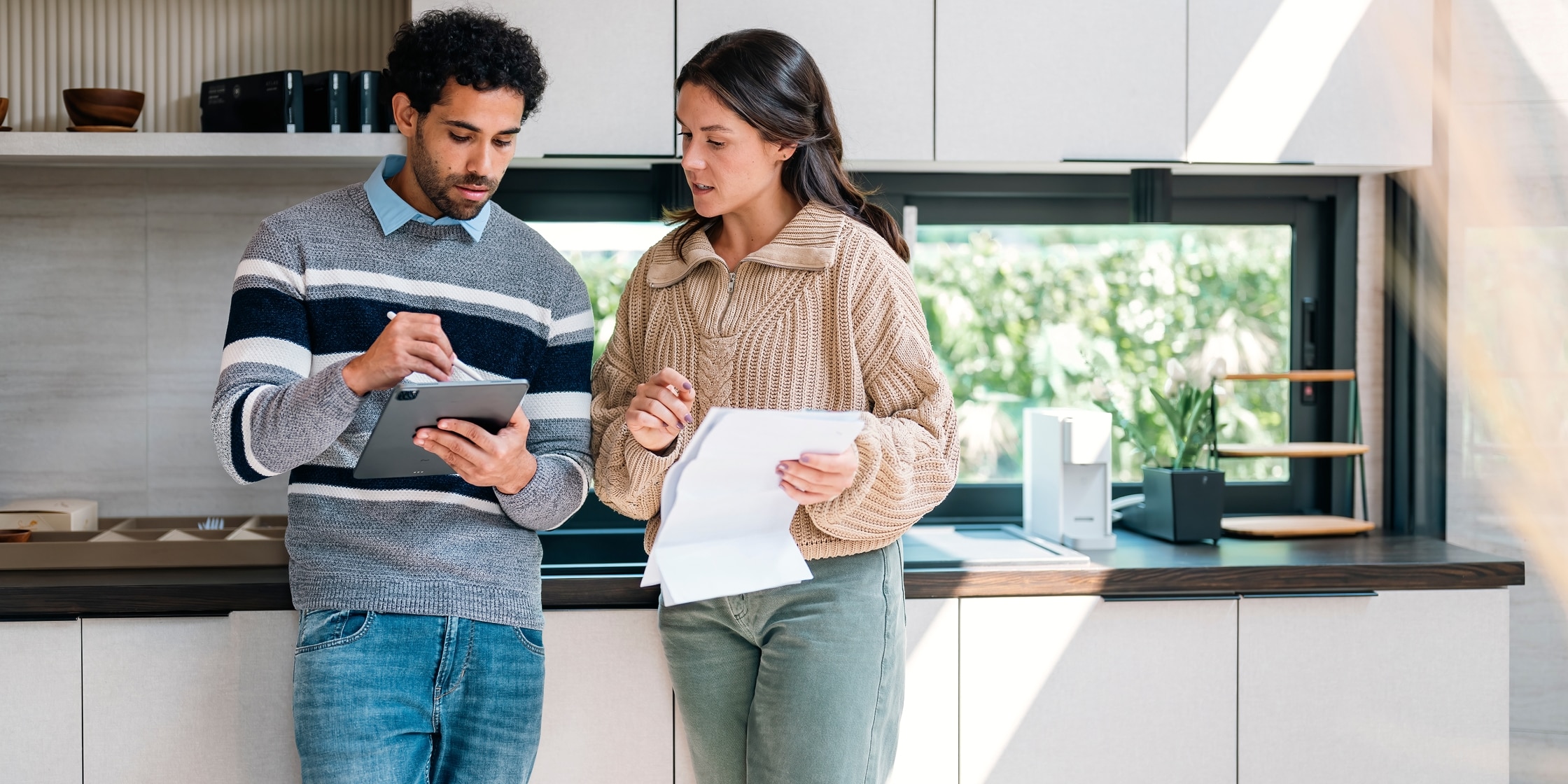 A man and woman review paperwork together in their kitchen while he uses a stylus on his tablet.