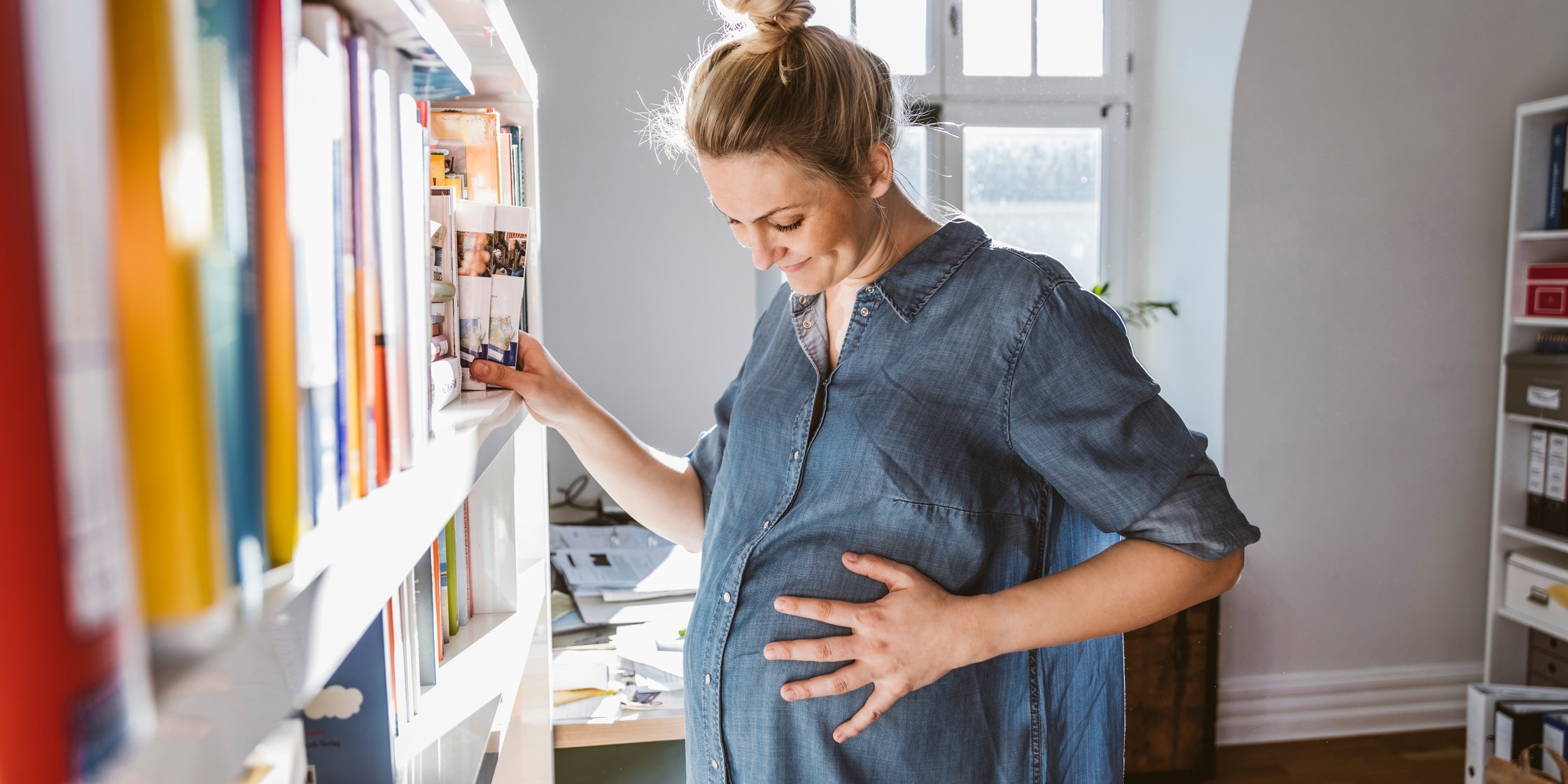 A pregnant woman looks lovingly down at her stomach while placing books on a bookshelf.