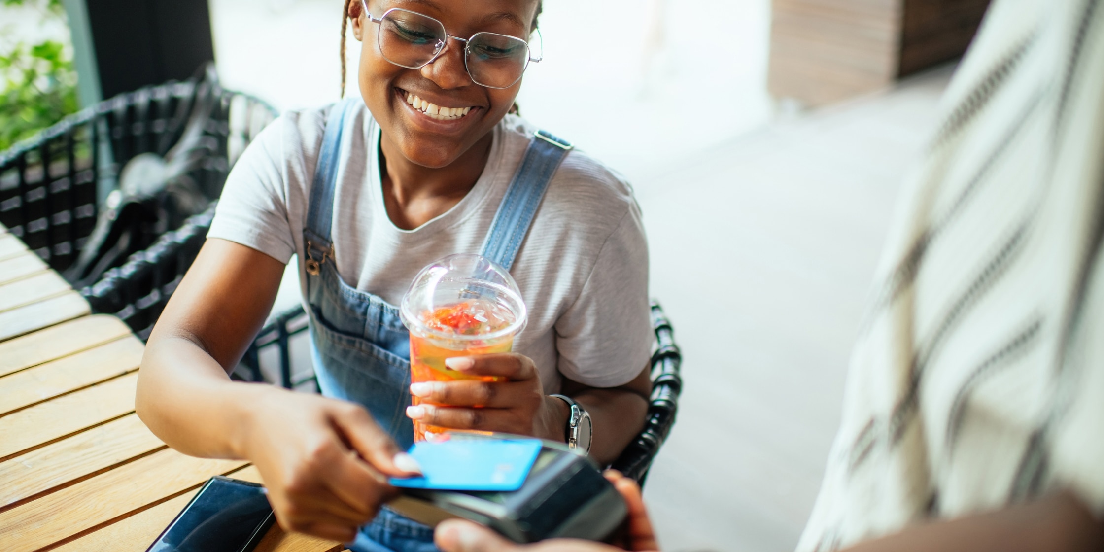 A young woman holding a brightly colored to-go beverage taps her credit card to pay.