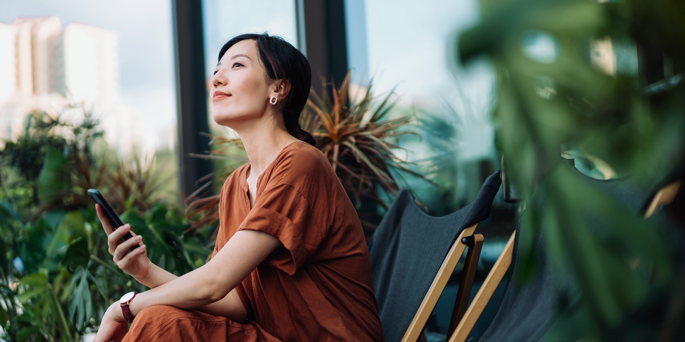 A woman outside holding her phone looks to the sky while sitting.