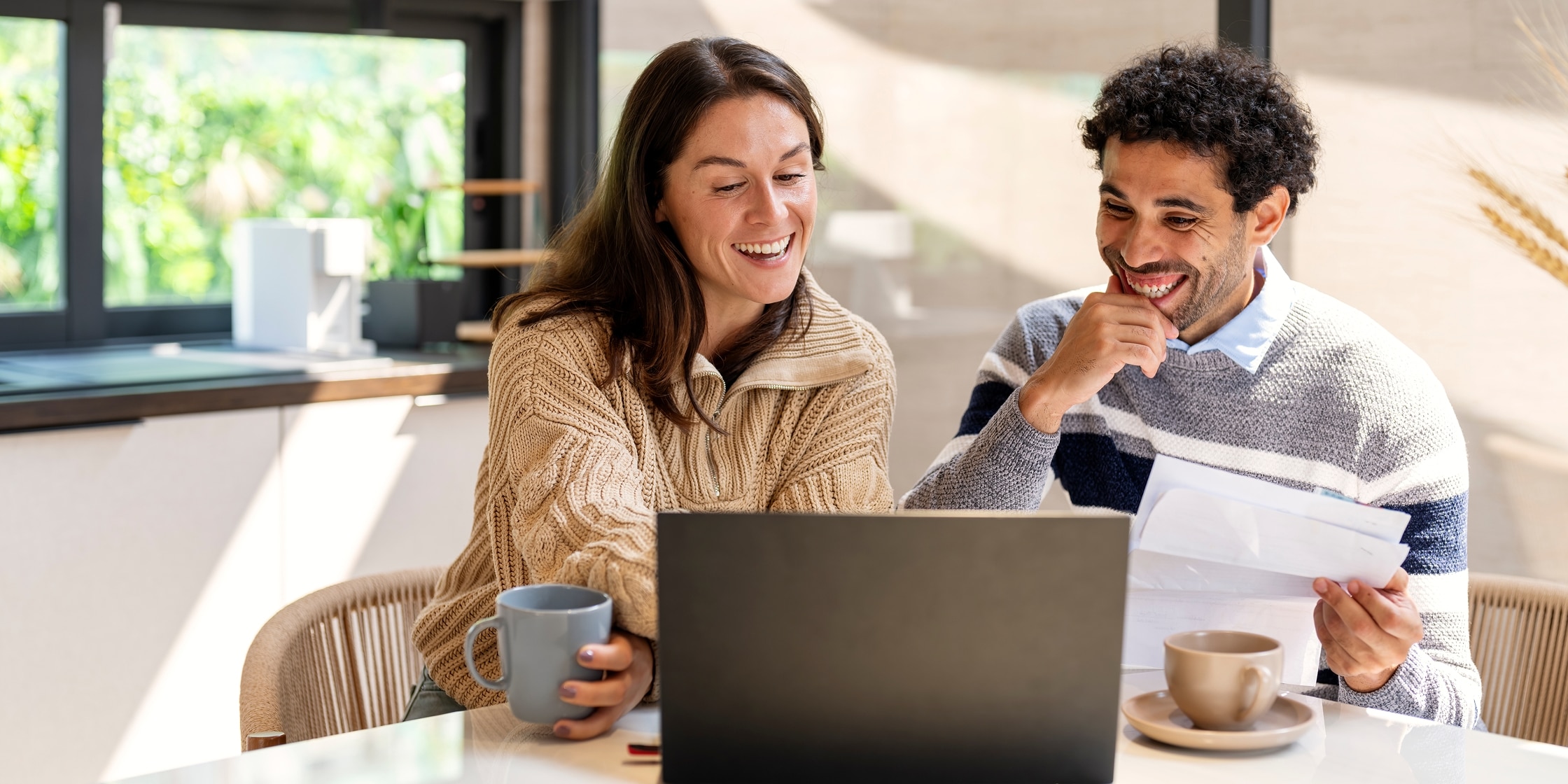 A couple sits at their kitchen table smiling and laughing together over morning coffee while looking at their laptop.