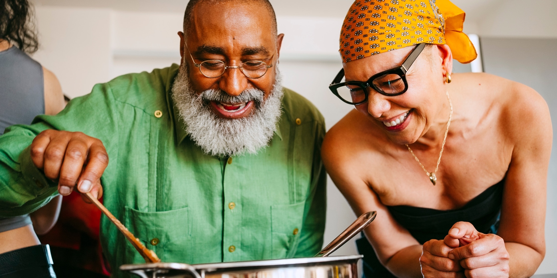 A man and woman cook together, stirring a large pot, while smiling and laughing.