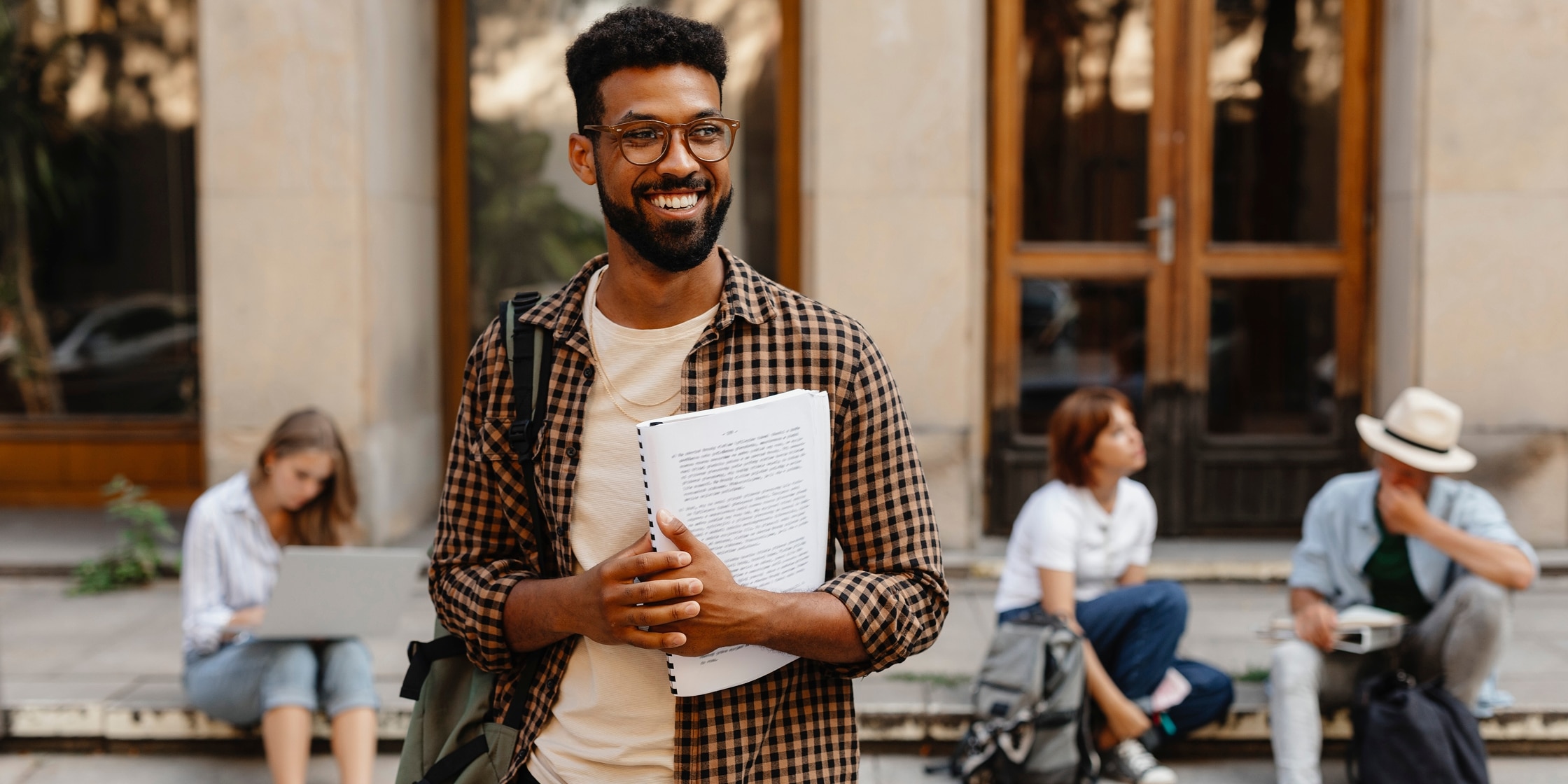 A smiling man grips a comb-bound booklet while the people behind him work and chat on the curb.