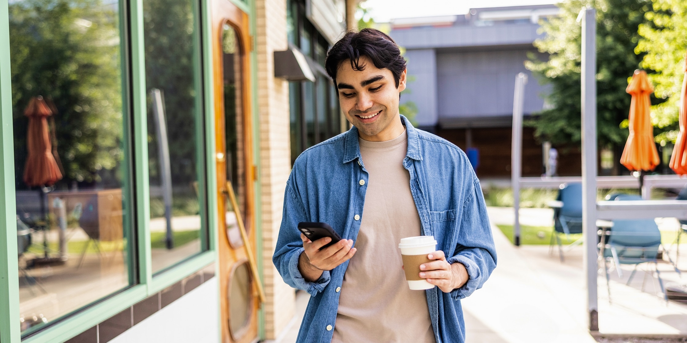 A young man smiles down at his smartphone while carrying his to-go cup of coffee.