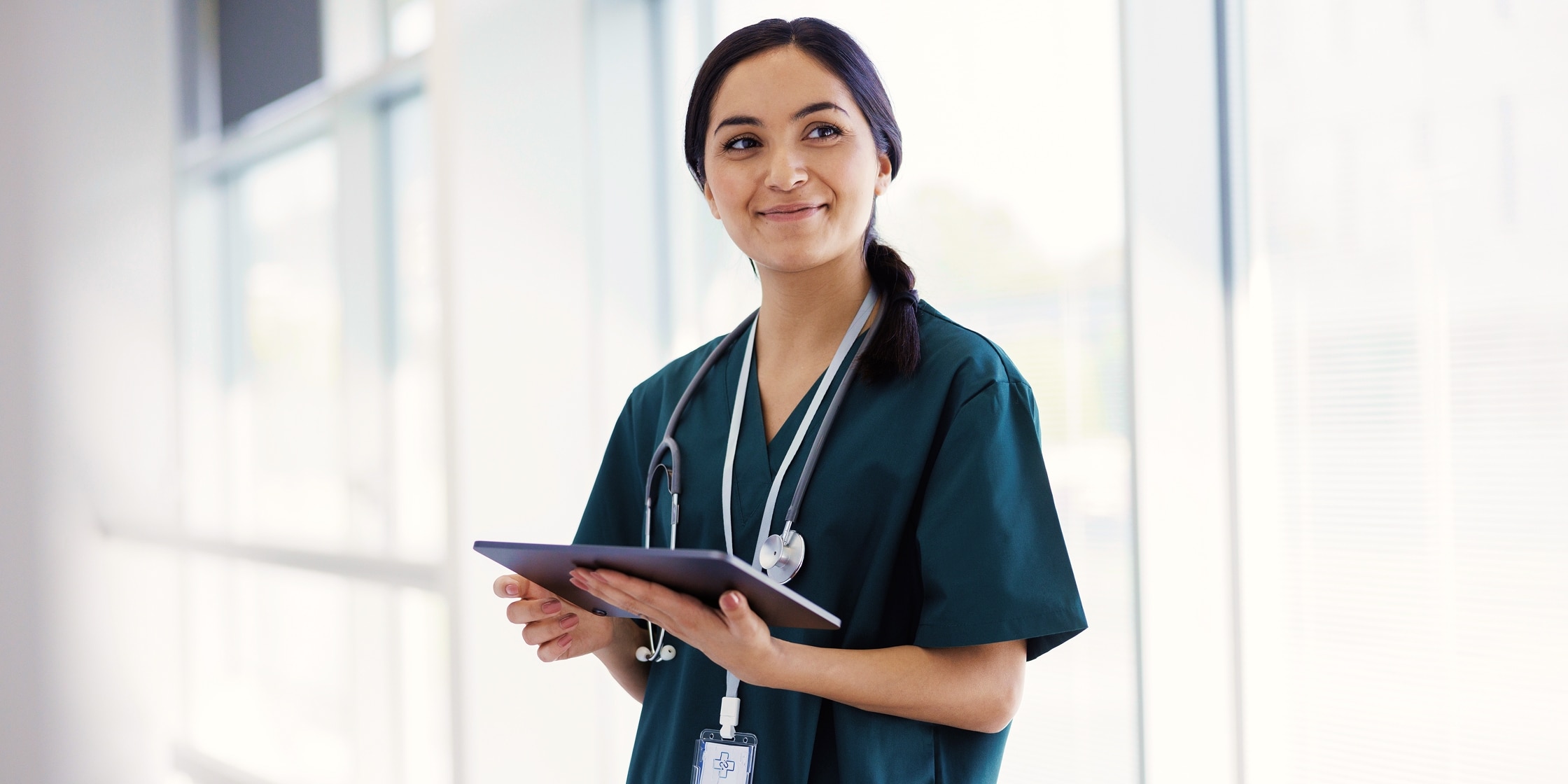 A medical professional looks off into the distance smiling and holding her tablet.