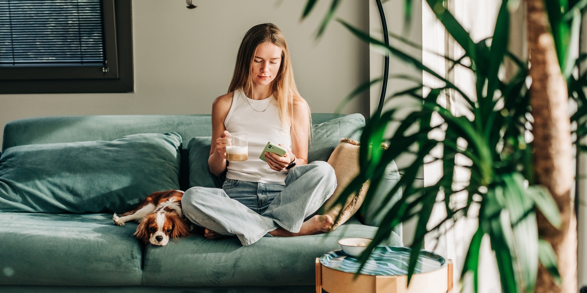 A young woman sits on her couch with her small dog drinking her morning coffee and looking at her smartphone.