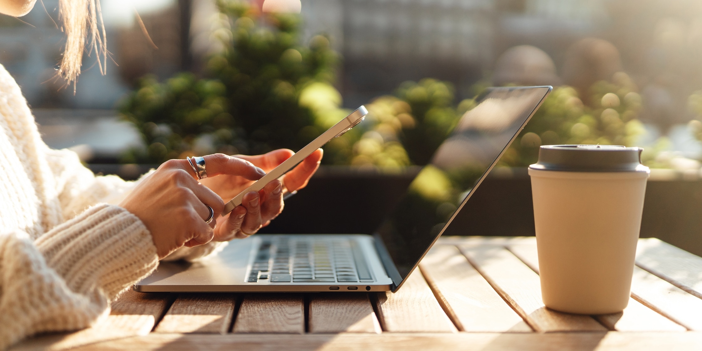 A woman uses her smartphone while her laptop and travel coffee cut sit in front of her on a small patio table.
