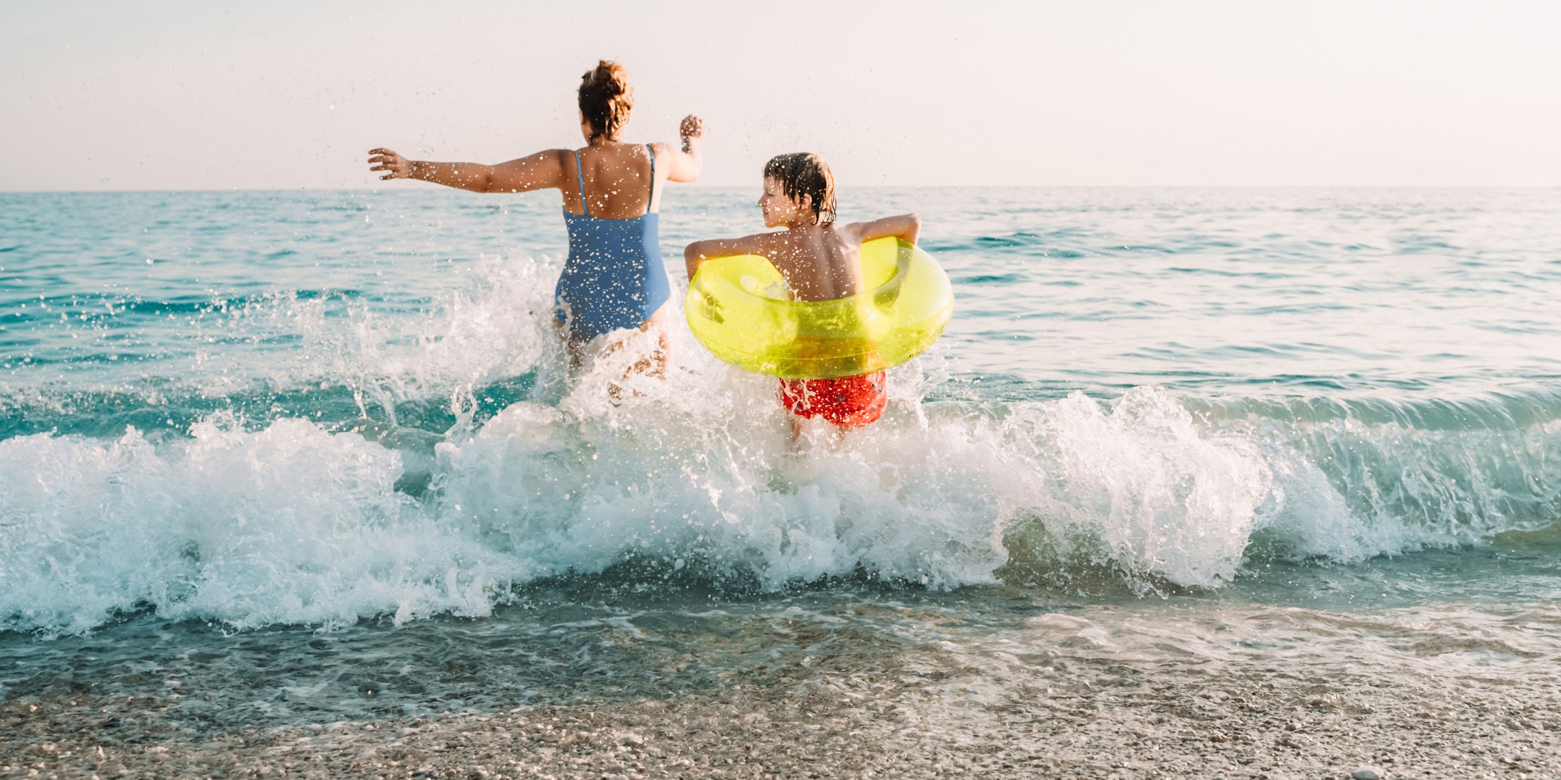 A woman and young child in a yellow inner tube splash into beach waves.
