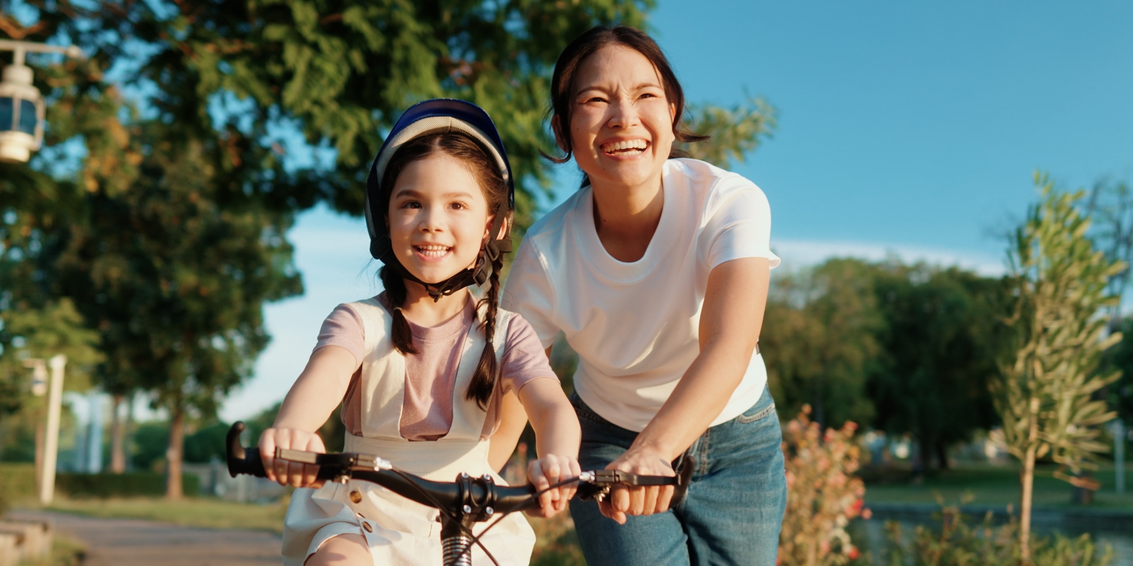 A woman happily steers a young girl learning to ride her bike in the park.