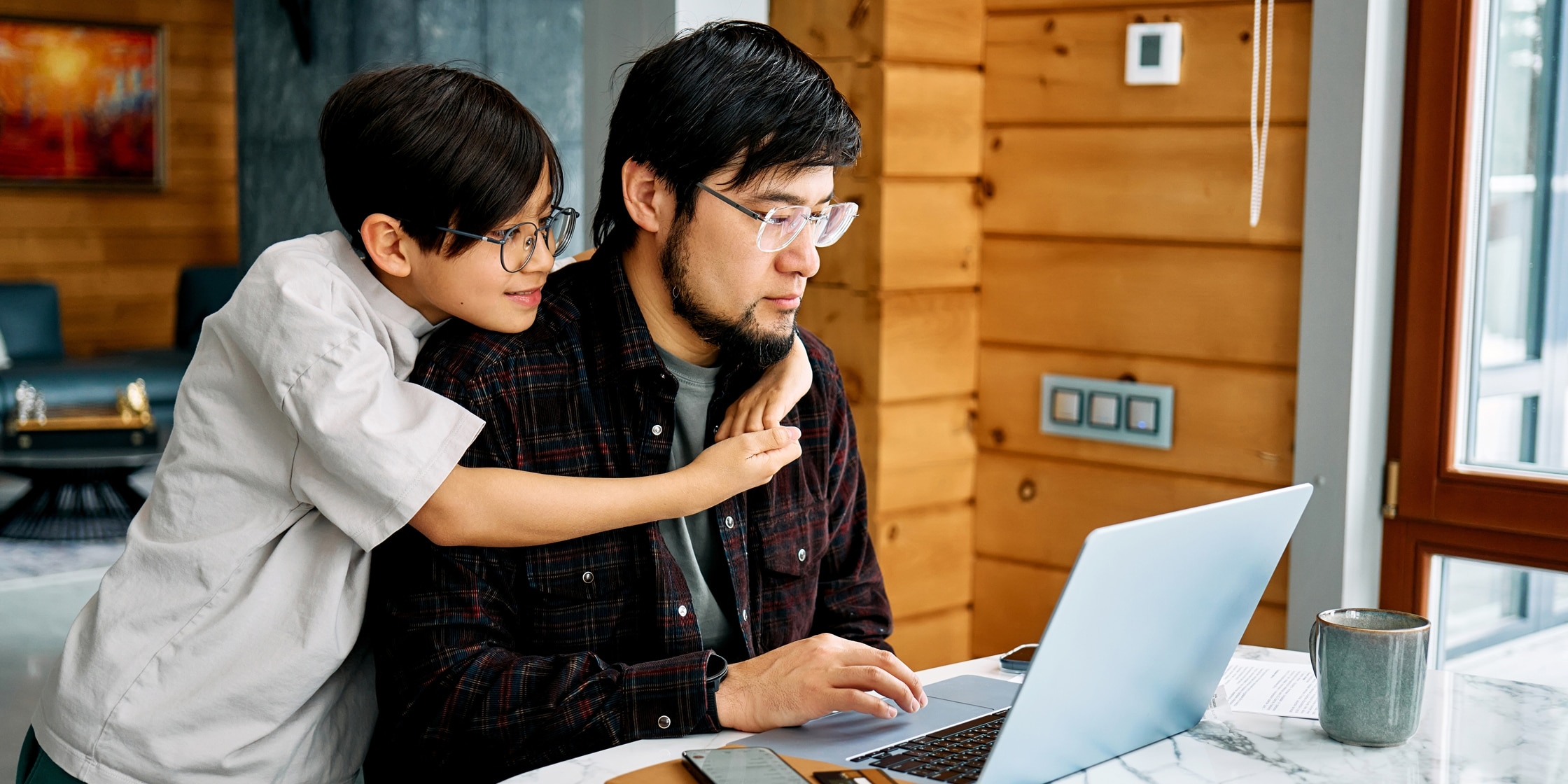 A child hugs his father’s shoulders while he types on a laptop at his kitchen table.