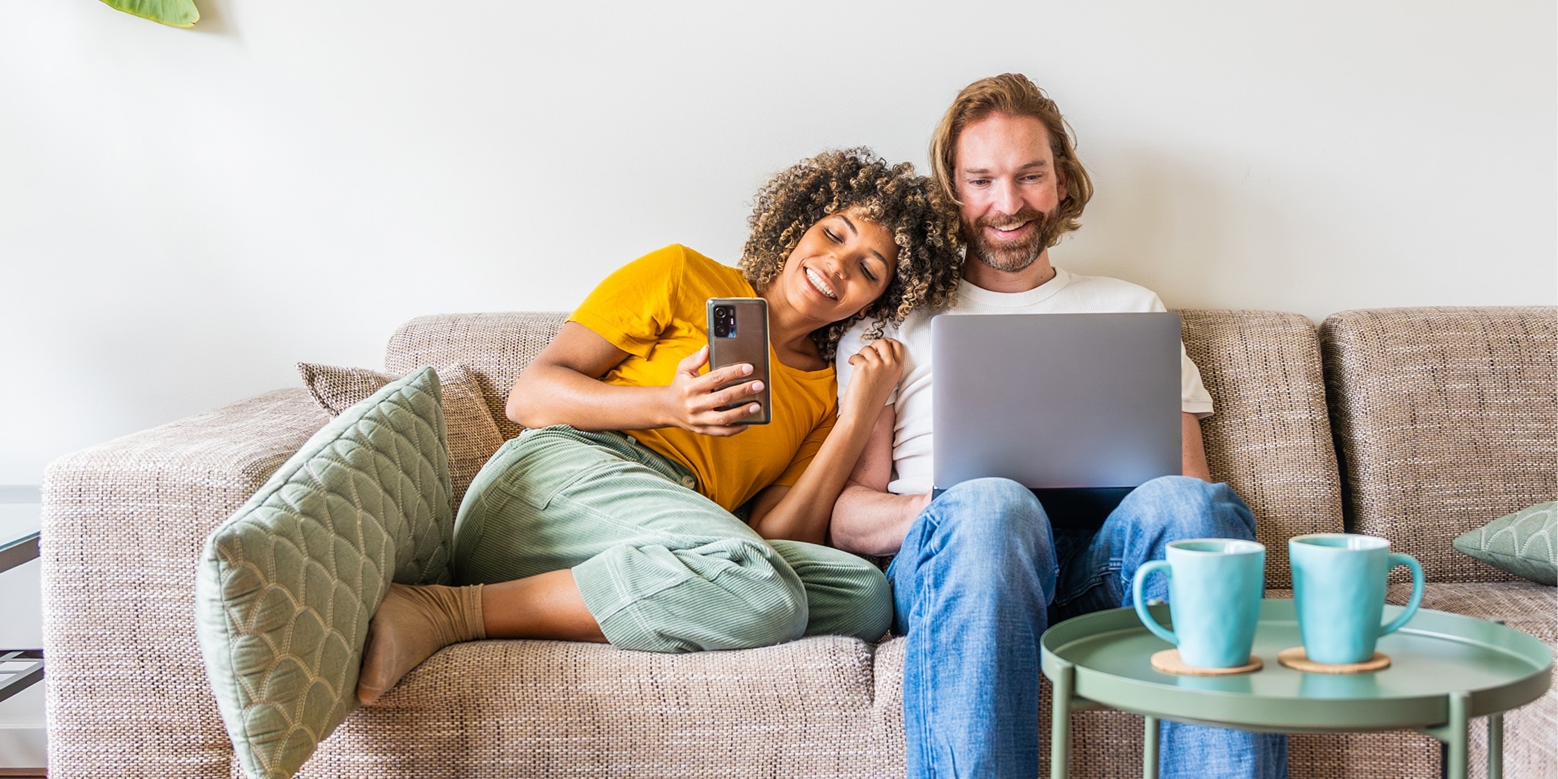 A couple sits on the couch together. She looks at her smartphone and he types on his laptop.
