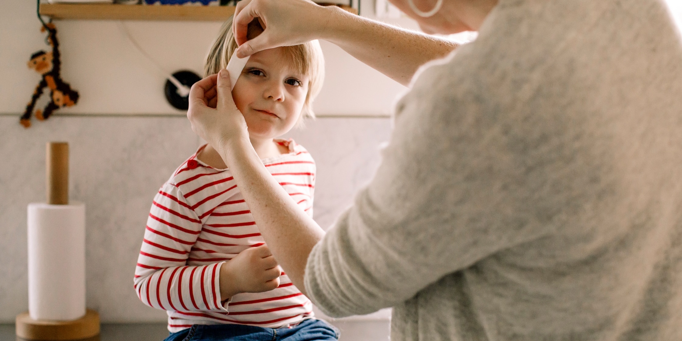 An adult places a bandage on the side of a young child’s face while he sits atop the kitchen counter.