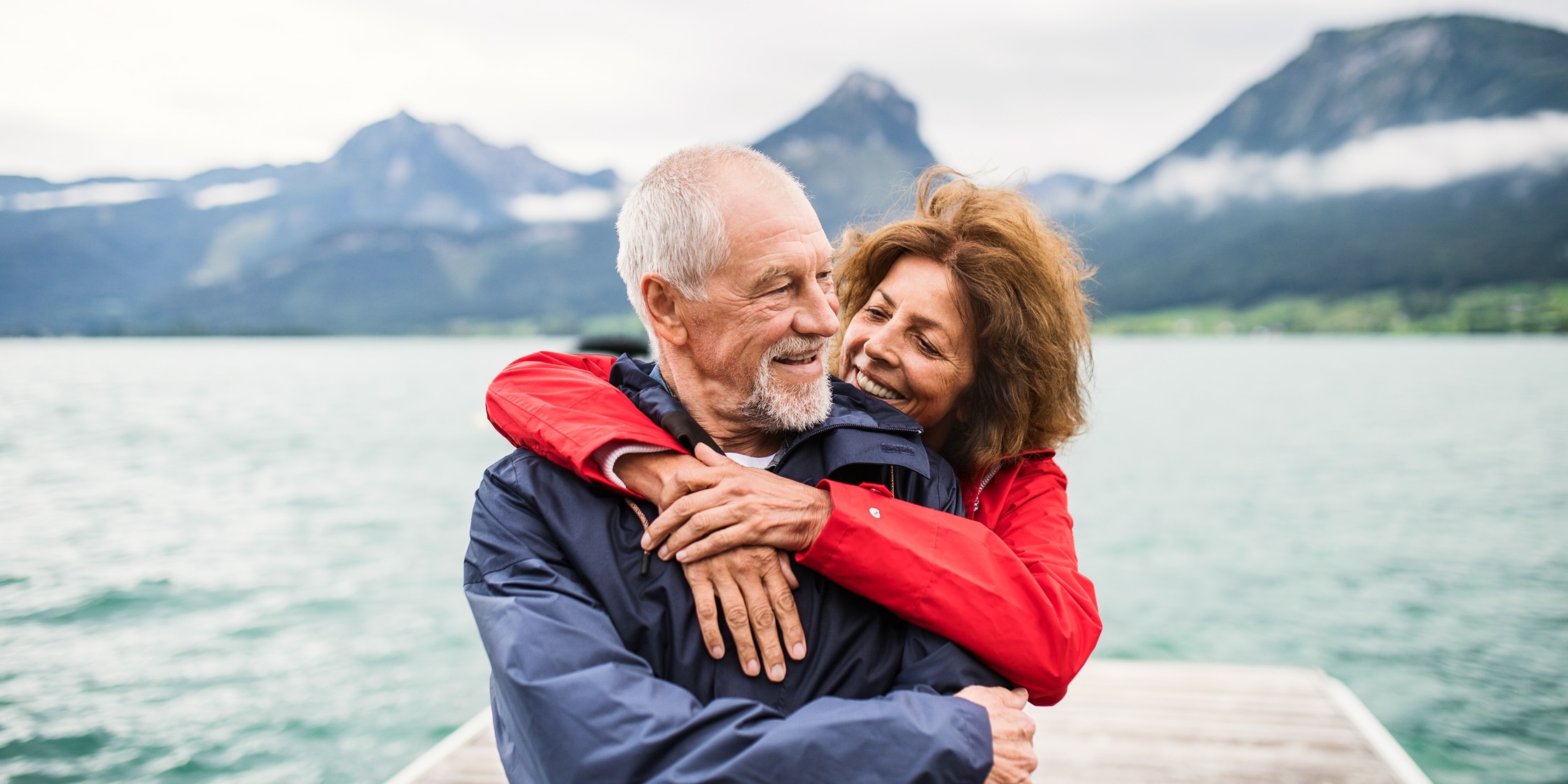 A senior couple embraces on a pier against the backdrop of a lake and mountain vista.
