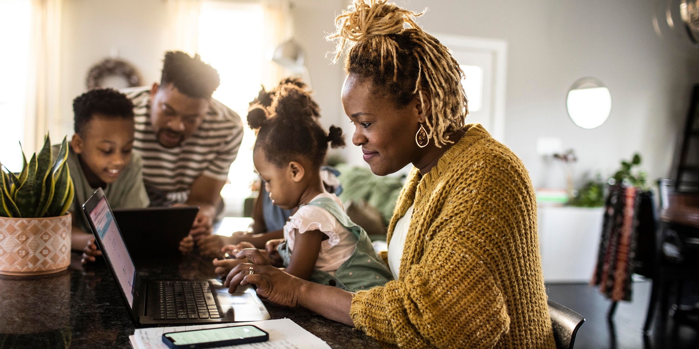 A couple and their two young children sit around the kitchen table using their tablet and laptop.